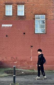Teenaged boy, dressed in black, on a skateboard, against the background of a red-brick wall.