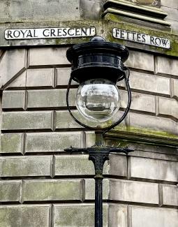 Moss-covered New Town masonry with an old fashioned lamppost reflecting King George V Park. Street-name signs for Royal Crescent and Fettes Row in the background.