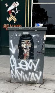Street art (woman's face) on utility box. Empty Red Bull can on top. Graffiti phrase 'Flew my way' underneath face.  
